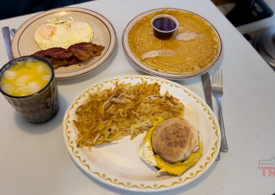 A hearty breakfast spread at Diana’s Diner in Albert Lea, MN, featuring eggs, crispy bacon, golden hash browns, a cheese and egg muffin, fluffy pancakes with syrup, and a glass of fresh orange juice on a white table.