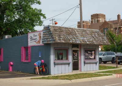 A small diner with pink trim and a shingled roof sits on a street corner in Albert Lea MN. A person in a pink helmet leans over a bicycle near the entrance of Diana’s Diner. Cars and a church are visible in the background.