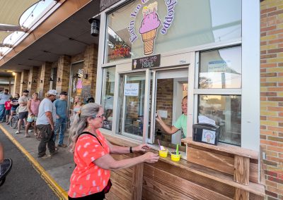 A woman receives ice cream at an outdoor pick-up window in Albert Lea, MN, while a line of people waits nearby. The stand has a sign with an ice cream cone above the window, with a brick building and shaded awnings in the background.