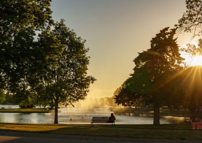 A person sits on a bench by Fountain Lake with a fountain at sunset, surrounded by trees in Albert Lea, MN. Sunlight filters through the branches, creating a peaceful atmosphere. The word TRAILS appears in red at the lower right.