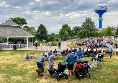 A crowd gathers for a summer event at a park in Albert Lea MN, with a gazebo, strollers, and people seated on the grass. The Albert Lea water tower and trees are visible in the background under a partly cloudy sky, capturing the lively spirit of Eddie Cochran Days.