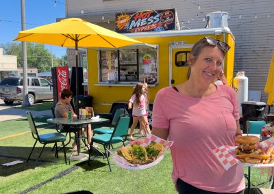 A woman smiles holding plates of food near the Hot Mess Express yellow food truck in Albert Lea, MN. People sit at outdoor tables under yellow umbrellas, enjoying the sunny and bright weather.