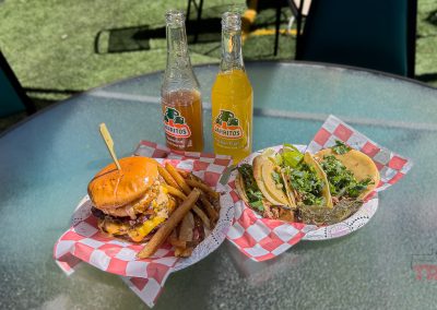 A table with a cheeseburger, fries, tacos, and two Jarritos sodas on a sunny outdoor patio at Albert Lea-Hot Mess Express. Food is served in red-and-white checkered paper baskets.