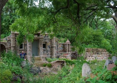 A small stone and rock building surrounded by lush green trees and plants; a man and child stand near the entrance, partially hidden by foliage, evoking the tranquil charm of the Itasca Rock Garden near Albert Lea MN.