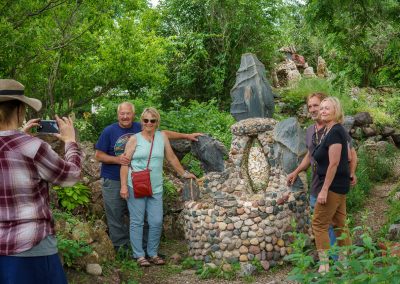 Five people pose by a stone sculpture in the lush, green Itasca Rock Garden in Albert Lea MN, while another person takes their photo. The group stands on a stone path surrounded by trees and plants.