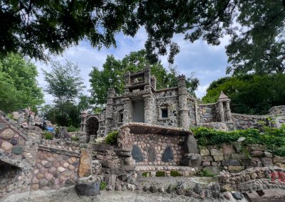 A stone castle-like structure with towers, surrounded by lush green trees and stone pathways under a partly cloudy sky, evokes the enchanting charm of the Itasca Rock Garden near Albert Lea MN.