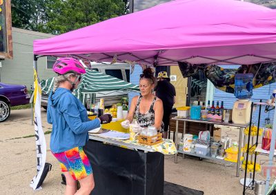 A cyclist in colorful gear buys a drink from a woman at an outdoor stand under a pink canopy in Albert Lea MN. The stand has various jars, cups, and equipment, while a car and tents are visible in the background.