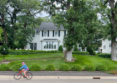 A person in a pink helmet rides a bike past a large white house with a well-manicured lawn, trees, and shrubs on a cloudy day in Albert Lea MN.
