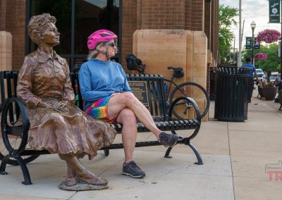 A person in a pink bike helmet sits on a bench next to the Marion Ross Statue, with a bicycle parked nearby on a city sidewalk in Albert Lea, MN. Shops and flowers are visible in the background.