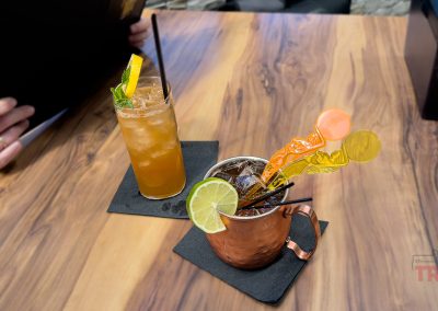 Two cocktails on a wooden table at the Thirsty Fox Bar in Albert Lea MN: one served in a copper mug with lime, orange stirrers, and ice, the other in a tall glass garnished with mint and lemon. In the background, a person holds a menu, ready to choose their next drink.