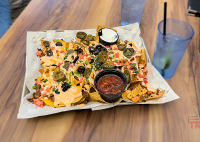 A tray of loaded nachos topped with cheese, olives, jalapeños, tomatoes, green onions, and served with salsa and sour cream sits on a wooden table at the Thirsty Fox Bar in Albert Lea MN, alongside two refreshing drinks.