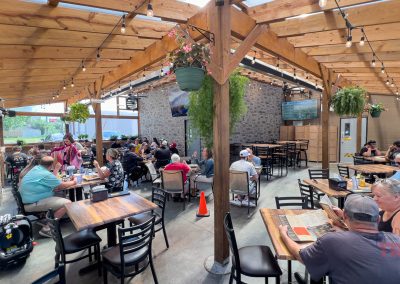 People dining at wooden tables under a covered patio at Thirsty Fox Bar in Albert Lea, with hanging plants, string lights, and TVs on the walls. The area is bright with natural sunlight, and most tables are occupied by groups of people eating and talking.