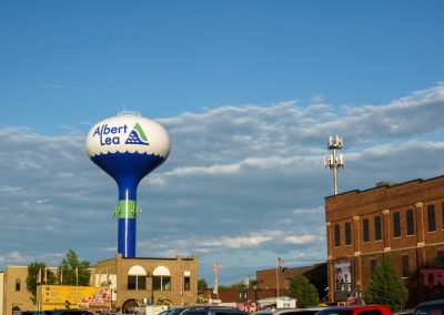 A large water tower labeled Albert Lea stands over brick buildings and parked cars under a blue sky with scattered clouds.