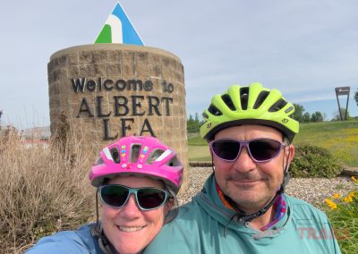 Two people wearing bike helmets and sunglasses smile in front of a large Welcome to Albert Lea stone sign on a sunny day with greenery and blue sky in the background.