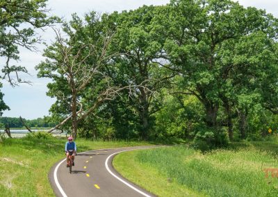 A person rides a bicycle on the Blazing Star Trail, a winding paved path in Minnesota surrounded by lush green trees and grass under a partly cloudy sky.