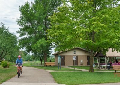 A person wearing a helmet rides a bicycle on a paved path in a green park along the Blazing Star Trail in Minnesota, passing a small building and picnic shelter with benches and tables, surrounded by trees.