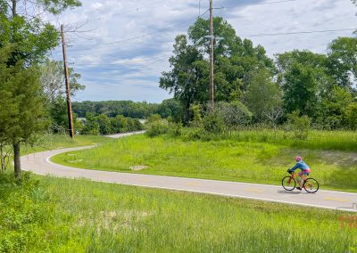 A person wearing a blue shirt and pink helmet rides a red bicycle on a paved, winding path through a green, grassy park under a partly cloudy sky along the Blazing Star Trail in Minnesota.