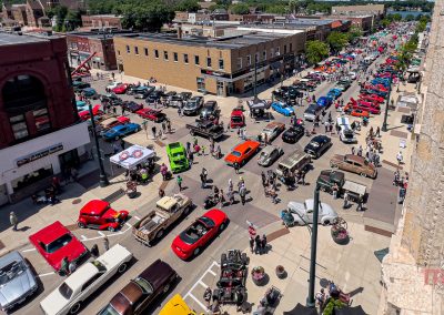 A busy downtown car show during Eddie Cochran Days in Albert Lea MN features colorful classic cars lining the streets, with many people walking around and admiring the vehicles on a sunny day.