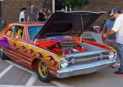 A vintage car with colorful purple and yellow flame paint is parked with its hood open at Eddie Cochran Days in Albert Lea, MN. Two men stand beside it, inspecting the engine at this lively outdoor car show, while other people and cars are visible in the background.
