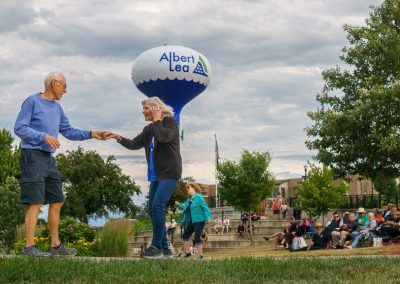 Two older adults dance on grass near a crowd seated outdoors during Eddie Cochran Days in Albert Lea MN. A water tower labeled Albert Lea is visible in the background under a cloudy sky. Trees and more people are in the scene, suggesting a lively community event.