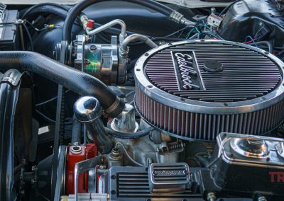 Close-up of a car engine bay featuring an Edelbrock air filter, various hoses, wires, and engine components—captured during Eddie Cochran Days in Albert Lea, MN. The image is detailed and shows the TRAILS logo in the bottom right corner.