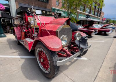 A vintage red fire truck is parked on a city street during Eddie Cochran Days in Albert Lea, MN, surrounded by other classic cars and shops.