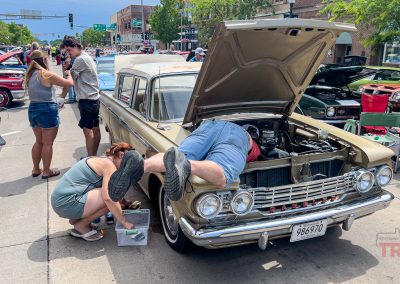 At Eddie Cochran Days in Albert Lea, MN, a person works under the hood of a vintage car while another kneels nearby. Classic cars line the street as people walk and observe, with trees providing shade on a sunny day.