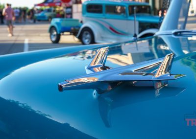 Close-up of a vintage car’s hood ornament shaped like a jet, with another classic car and people blurred in the background at an outdoor car show during Eddie Cochran Days in Albert Lea MN.
