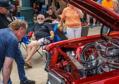 A man leans in to look closely at a shiny red classic car engine at an outdoor car show during Eddie Cochran Days in Albert Lea, MN, while people sit and stand around, observing and relaxing.