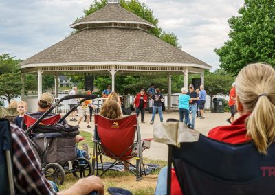 People enjoy live music at a park gazebo during Eddie Cochran Days in Albert Lea, MN; some are seated in chairs while others stand or dance. Children and adults are present, surrounded by trees and a cloudy sky.
