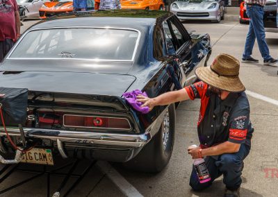 A person in a straw hat and racing shirt polishes the back of a black classic car at a car show during Eddie Cochran Days in Albert Lea MN. Several other people and colorful cars are visible in the background.