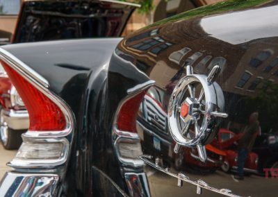 Close-up of a classic black car’s rear fin and emblem, reflecting nearby buildings and a red vintage car with its hood open at an outdoor car show during Eddie Cochran Days in Albert Lea, MN. The word TRAILS appears in red on the bottom right.