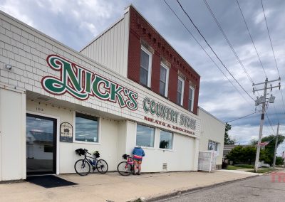 Two bicycles are parked outside Nick’s Country Store, a classic white building with red accents in Hayward MN. The store sign advertises meats and groceries, while a cloudy sky looms overhead and power lines run alongside the street.