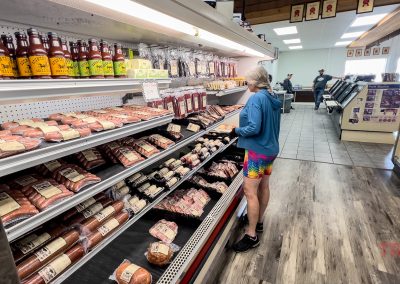 A person in colorful shorts browses packaged meats and sausages in a grocery store aisle at Nick’s Country Store in Hayward, MN. Shelves are stocked with various items, and another person is visible near the counter in the background.