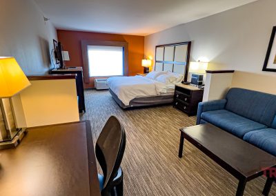 Experience comfort and convenience in this Holiday Inn Express hotel room in Albert Lea, MN. The space features a cozy bed, nightstands, lamps, blue sofa, coffee table, desk, chair, TV, and a window that fills the room with natural light. The white and orange walls are accented by a stylish painting above the sofa.