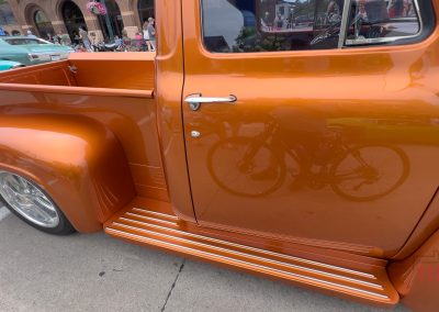 An orange vintage truck is parked at a car show during Eddie Cochran Days in Albert Lea, MN. The shiny door reflects a person riding a bicycle, while other classic cars and people are visible in the background.